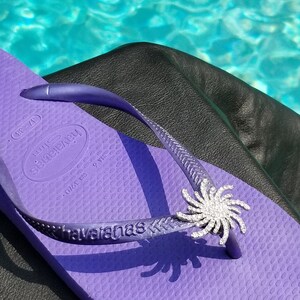 Op de afbeelding: Paarse flip-flop met een zilveren strass-bloemversiering. De flip-flop heeft de merknaam "havaianas" op de band gedrukt. De flip-flop ligt op een zwart oppervlak.