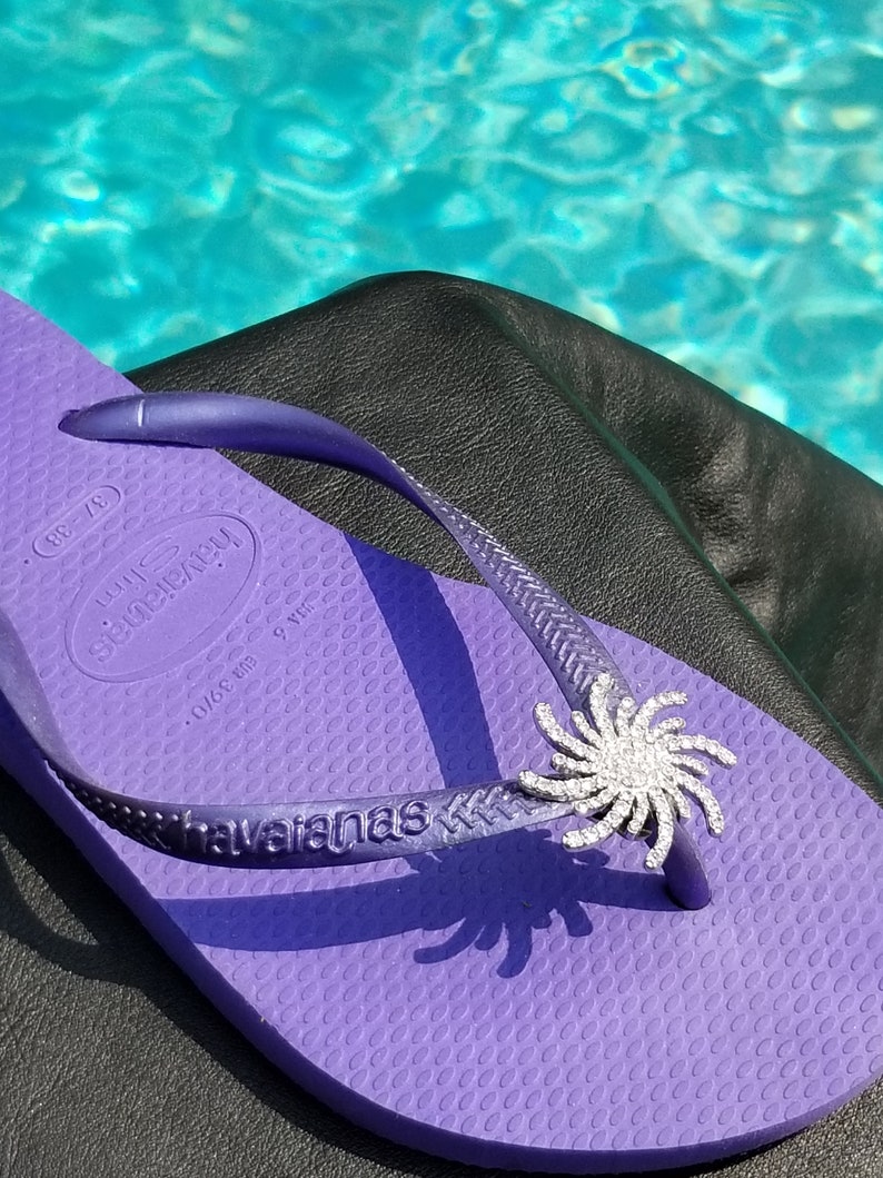 Op de afbeelding: Paarse flip-flop met een zilveren strass-bloemversiering. De flip-flop heeft de merknaam "havaianas" op de band gedrukt. De flip-flop ligt op een zwart oppervlak.