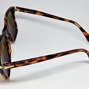 May include: Tortoiseshell sunglasses with brown tinted lenses and gold-tone metal accents at the hinges. The inner temple arms feature the text GIORGIO ARMANI and MADE IN ITALY, while the outer temple arm displays the GIORGIO ARMANI brand name.