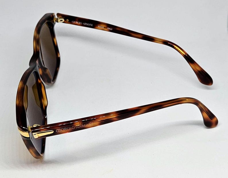 May include: Tortoiseshell sunglasses with brown tinted lenses and gold-tone metal accents at the hinges. The inner temple arms feature the text GIORGIO ARMANI and MADE IN ITALY, while the outer temple arm displays the GIORGIO ARMANI brand name.