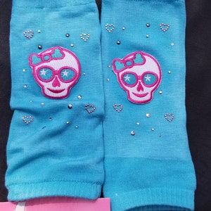 May include: A pair of turquoise blue leg warmers with a pink skull and crossbones design. The skull has pink sunglasses and a pink bow. The leg warmers are decorated with silver glitter and rhinestones. The leg warmers are labeled "infants sizes 6-24m Legwarmers".