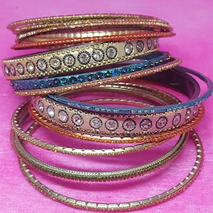 May include: A stack of colorful bangles with various designs. Some bangles are gold-toned with small, sparkling embellishments. Others feature cream-colored bands with clear, round rhinestones. The background is a solid pink.