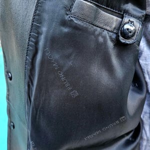 Pu&ograve; includere: L'interno di una giacca in pelle nera caratterizzata da una fodera nera liscia con il testo ripetuto BRUNO MAGLI. La giacca include una tasca interna singola con bordo in pelle e chiusura con bottone nero, oltre a bottoni neri visibili.