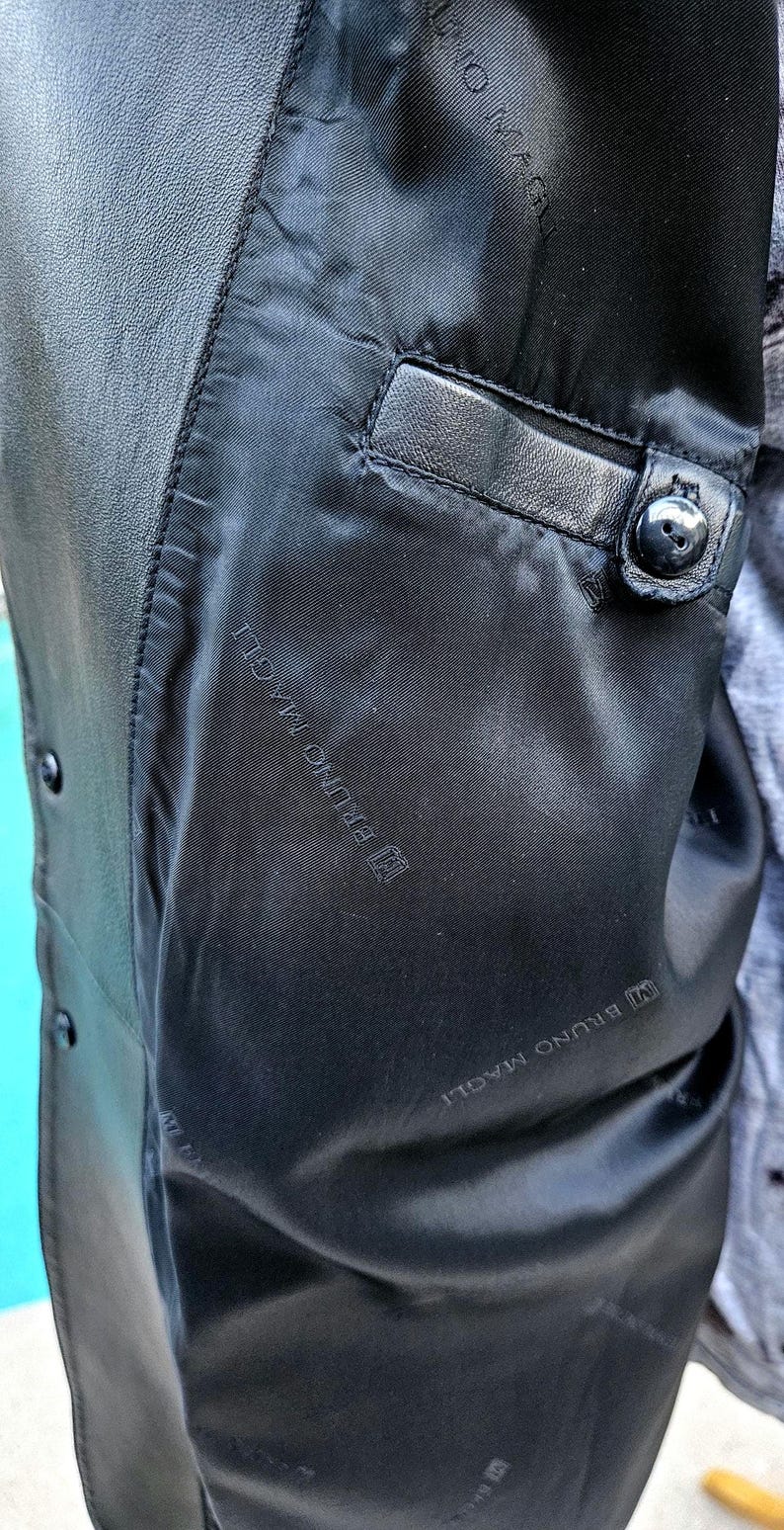 Pu&ograve; includere: L'interno di una giacca in pelle nera caratterizzata da una fodera nera liscia con il testo ripetuto BRUNO MAGLI. La giacca include una tasca interna singola con bordo in pelle e chiusura con bottone nero, oltre a bottoni neri visibili.