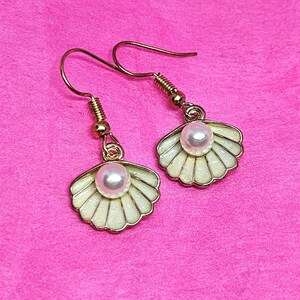 May include: A pair of gold-colored seashell earrings with a pearl in the center of each shell. The shells are off-white with gold trim and hang from gold-colored ear wires. The earrings are displayed on a pink background.