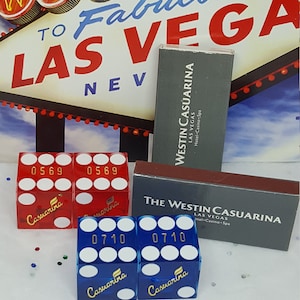 May include: Two red dice with the number 0569 and the word "Casuarina" on them, and two blue dice with the number 0710 and the word "Casuarina" on them. The dice are on a white surface. There are two matchboxes with the text "The Westin Casuarina Las Vegas Hotel Casino Spa" on them.