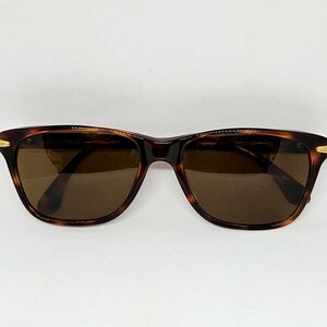 May include: Tortoiseshell-patterned sunglasses with brown lenses and gold-tone metal accents at the hinges. The inner frame features the text Giorgio Armani Made in Italy. These sunglasses have a classic rectangular shape with rounded edges.