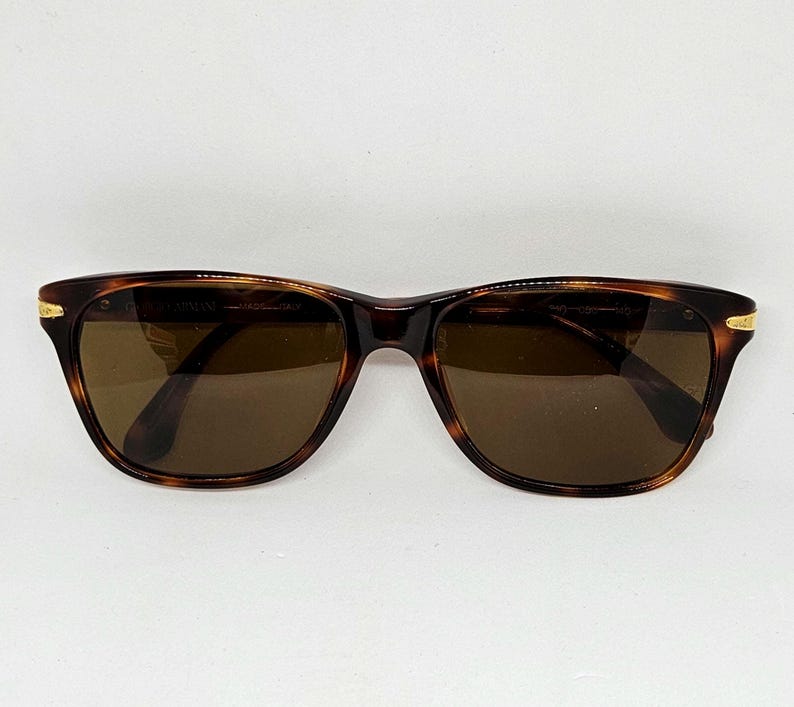 May include: Tortoiseshell-patterned sunglasses with brown lenses and gold-tone metal accents at the hinges. The inner frame features the text Giorgio Armani Made in Italy. These sunglasses have a classic rectangular shape with rounded edges.