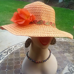 May include: A straw hat with a wide brim, orange flower, and embroidered trim. The hat is on a mannequin with a necklace and earrings.