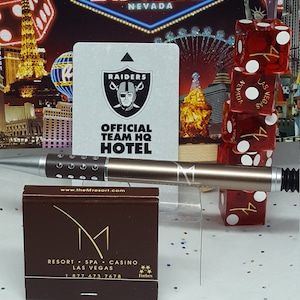 May include: A brown matchbook with a white 'M' logo and the text 'RESORT SPA CASINO LAS VEGAS 1 877 673 7678 Forbes' printed on it. The matchbook is sitting on a white surface with a silver pen and a stack of red dice with white dots.