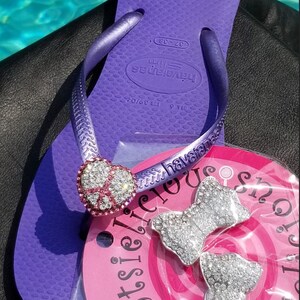 May include: Purple flip flops with a heart-shaped charm and rhinestones. The flip flops have the brand name "havaianas" printed on the strap. The flip flops are on a pink background with the text "delicious shop" printed on it. There are two silver bow charms with rhinestones on the pink background.