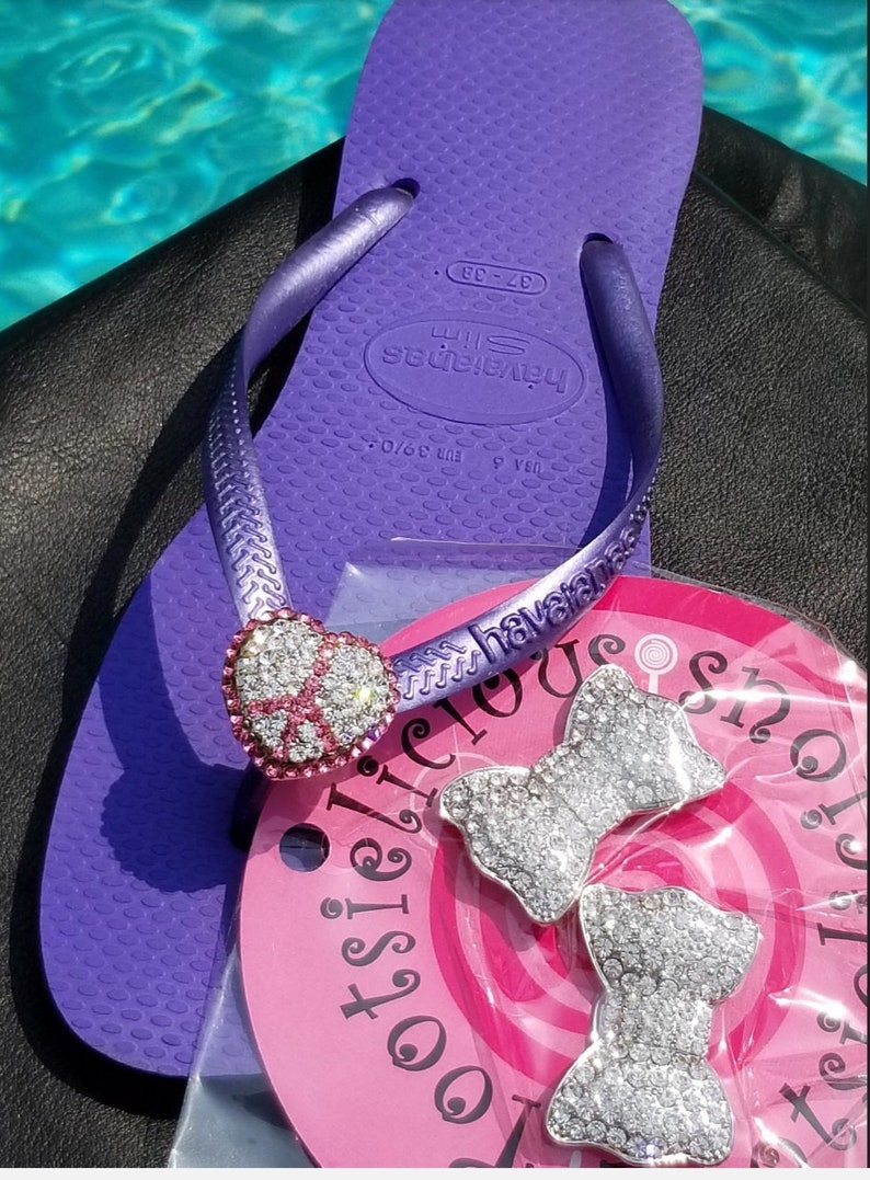 May include: Purple flip flops with a heart-shaped charm and rhinestones. The flip flops have the brand name "havaianas" printed on the strap. The flip flops are on a pink background with the text "delicious shop" printed on it. There are two silver bow charms with rhinestones on the pink background.