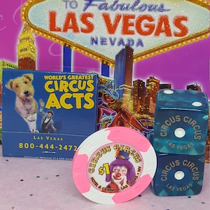 May include: A pink and white casino chip with a clown image and the text "Circus Circus Hotel & Casino Las Vegas, NV".  Two blue dice with the text "Circus Circus Las Vegas" are in the background. A box with the text "World's Greatest Circus Acts Las Vegas 800-444-2472" is in the lower left corner. The background is a pink and white glittery image of the "Welcome to Fabulous Las Vegas, Nevada" sign.