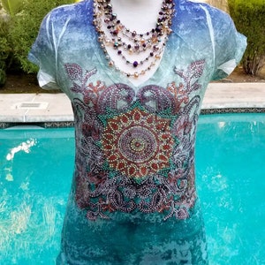 May include: A turquoise blue, white, and purple ombre t-shirt with a beaded mandala design. The shirt has a v-neck and short sleeves. The model is wearing a multi-strand necklace with purple, gold, and clear beads.