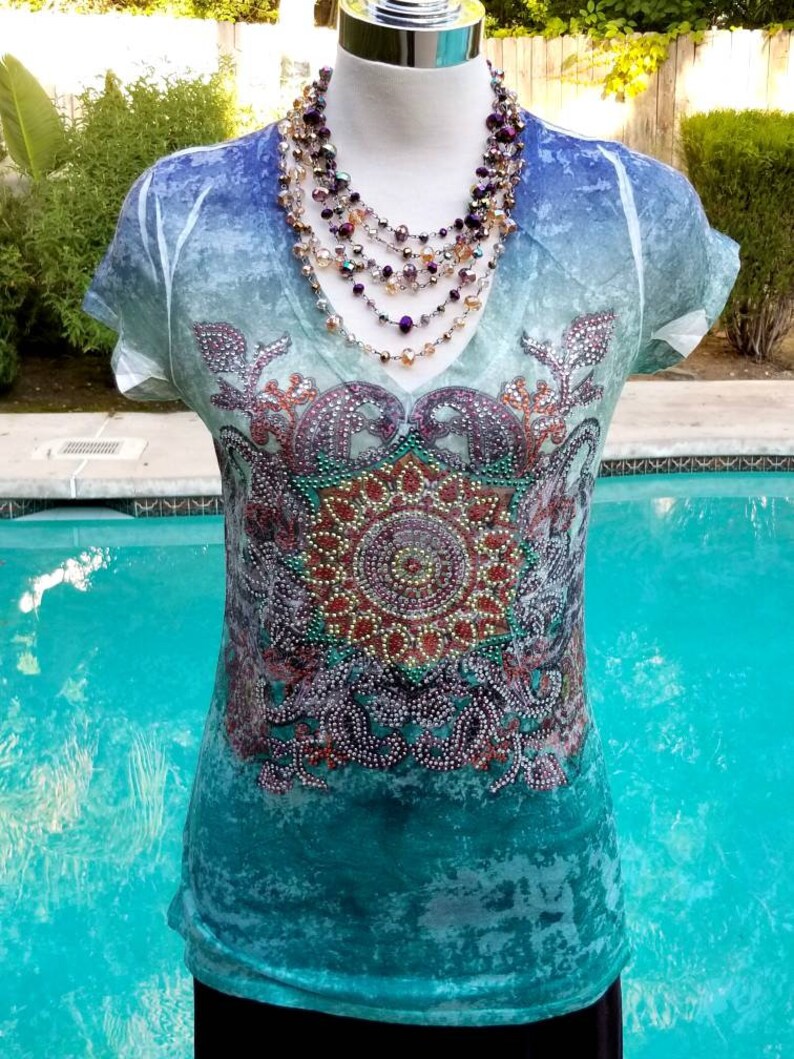 May include: A turquoise blue, white, and purple ombre t-shirt with a beaded mandala design. The shirt has a v-neck and short sleeves. The model is wearing a multi-strand necklace with purple, gold, and clear beads.