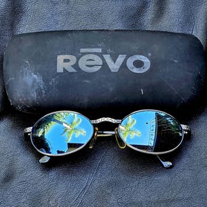 May include: Oval sunglasses with silver-toned frames featuring a textured pattern and reflective blue lenses. The glasses are positioned in front of a black, rectangular hard-shell carrying case with the brand name REVO printed in white letters.