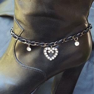 May include: Black leather boots with a silver chain ankle bracelet featuring a heart charm with rhinestones.