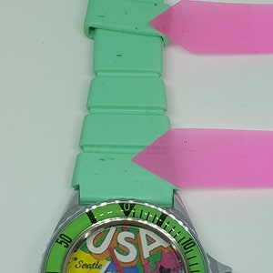 May include: A green and silver wristwatch with a map of the USA on the face. The watch has a green silicone band and a silver bezel with black markings. The watch face features the words "USA" and the names of several major cities.