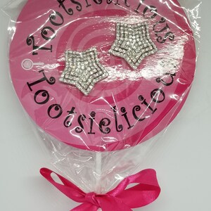 May include: A pink lollipop with a white stick and a pink ribbon bow. The lollipop has a white circle with black text that says "Lootsielicious". Two silver star-shaped earrings with rhinestones are attached to the lollipop.