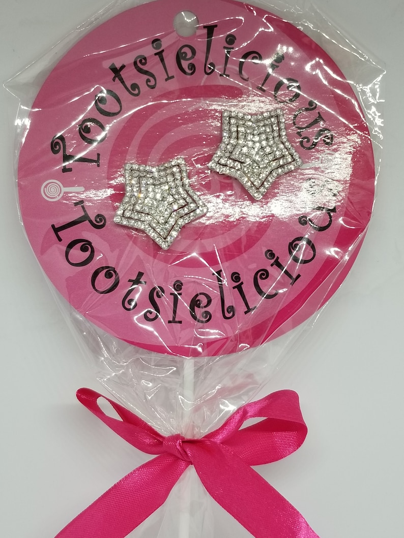May include: A pink lollipop with a white stick and a pink ribbon bow. The lollipop has a white circle with black text that says "Lootsielicious". Two silver star-shaped earrings with rhinestones are attached to the lollipop.