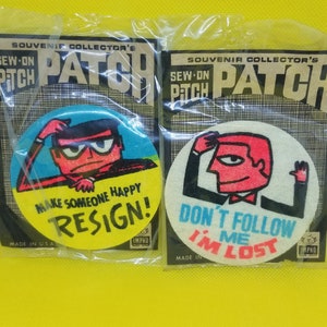 May include: Two round sew-on patches with cartoon characters and text. The first patch is yellow with a blue-haired character with a red face and the text "Make someone happy, Resign!" The second patch is white with a red-haired character with a red face and the text "Don't follow me, I'm lost!"