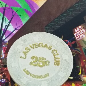 May include: A white casino chip with gold lettering. The chip says "Las Vegas Club" and "25c" in the center. The chip also says "Las Vegas, NV" around the edge.