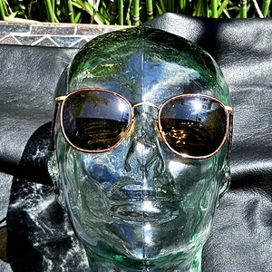 May include: A pair of sunglasses with round, dark brown lenses and a thin, metallic gold-toned frame. The sunglasses are displayed on a clear, glass-like mannequin head, showcasing the reflective surface of the lenses and the metallic bridge.