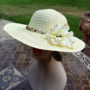 May include: A large, wide-brimmed straw hat with a white and gold flower accent. The hat is trimmed with a band of gold sequins.