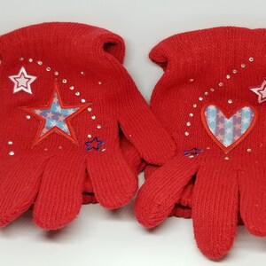 May include: Red knit gloves with a star and heart design. The star and heart are embroidered with red, white, and blue stripes. The gloves are decorated with rhinestones.