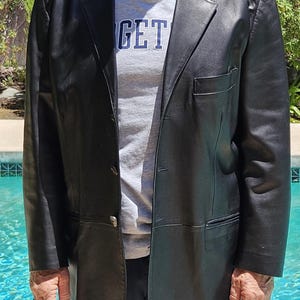 Pu&ograve; includere: Un blazer in pelle nera con revers dentellato, chiusura frontale a tre bottoni e una tasca singola sul petto. Il blazer &egrave; indossato sopra una t-shirt grigia con il testo parziale GET visibile in lettere blu scuro sulla parte anteriore.
