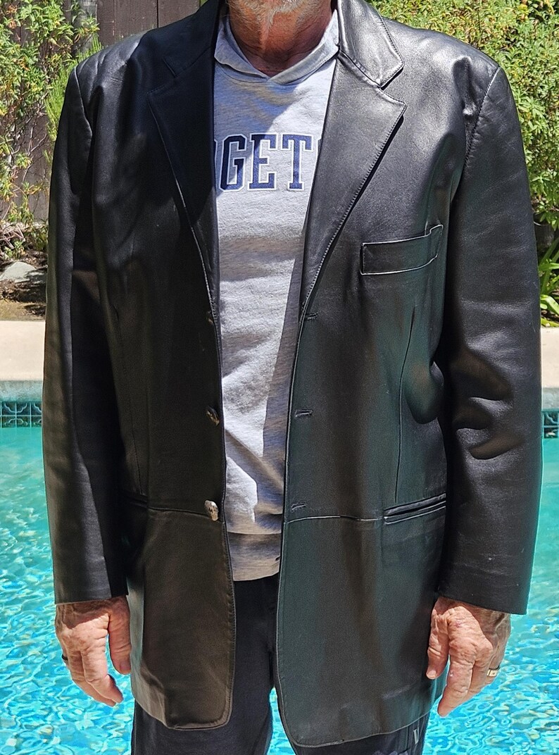 Pu&ograve; includere: Un blazer in pelle nera con revers dentellato, chiusura frontale a tre bottoni e una tasca singola sul petto. Il blazer &egrave; indossato sopra una t-shirt grigia con il testo parziale GET visibile in lettere blu scuro sulla parte anteriore.