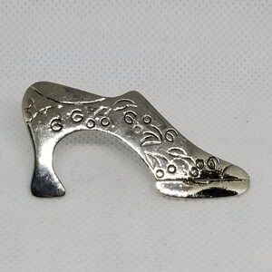 May include: Silver shoe charm with an intricate floral design. The charm is shaped like a high-heeled shoe with a pointed toe.