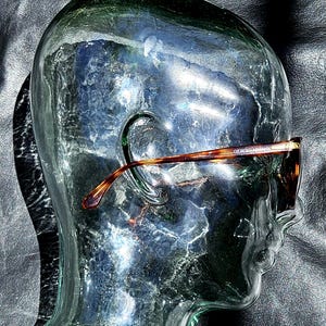May include: Tortoiseshell-patterned eyeglasses with a brown frame and thin temples, featuring the text Giorgio Armani printed on the side, displayed on a clear glass head sculpture that highlights the frame's curved design and color details.