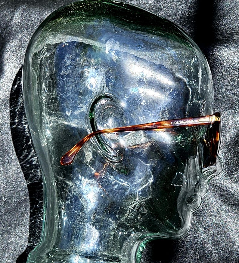 May include: Tortoiseshell-patterned eyeglasses with a brown frame and thin temples, featuring the text Giorgio Armani printed on the side, displayed on a clear glass head sculpture that highlights the frame's curved design and color details.