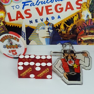 May include: A red and black casino chip with the text "Terrible's Town $5 Pahrump, Nevada" and a cartoon cowboy on it. Two red dice with white dots and the text "Terrible's Hotel Casino" are next to the chip. A clear keychain with a cartoon cowboy wearing a red vest and a black hat is in the background.