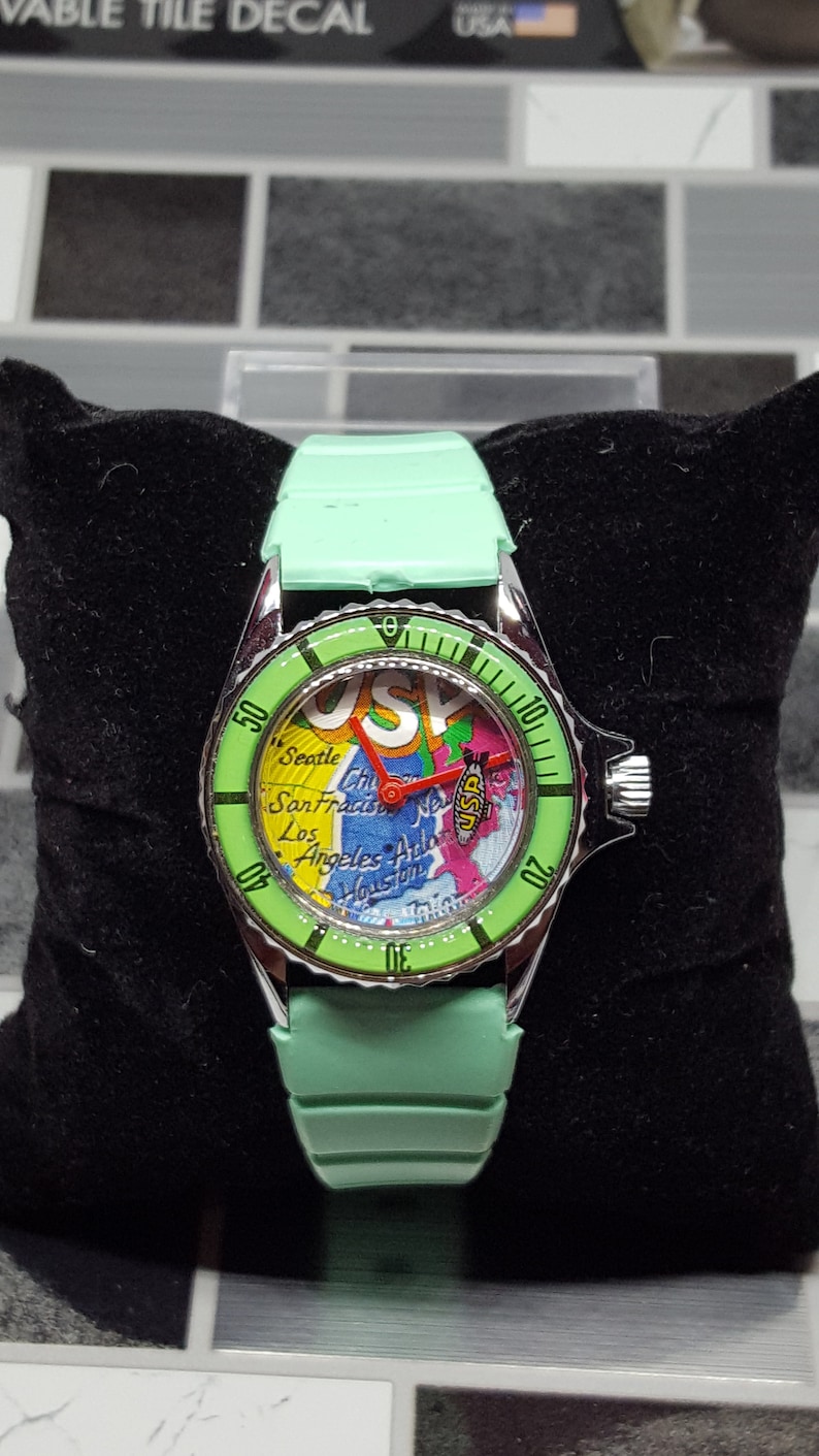 May include: A green wristwatch with a map of the USA on the face. The watch has a silver bezel with black markings and a green strap.