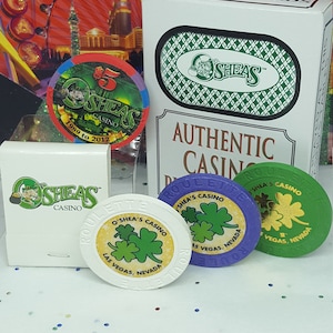 May include: A set of four O'Shea's Casino playing chips. The chips are green, purple, white, and yellow. The chips have a shamrock design and the text "O'Shea's Casino Las Vegas, Nevada". The chips are on a white surface with a box of playing cards in the background. The box is white with green and white checkered pattern and the text "Authentic Casino Playing Cards" and "O'Shea's".