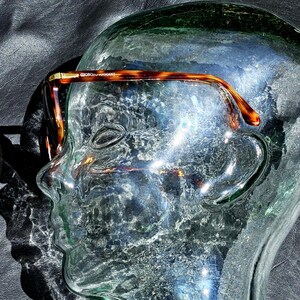May include: Tortoiseshell-patterned eyeglasses with a brown and gold frame. The temple arm features the text GIORGIO ARMANI in gold lettering. The glasses are positioned on a clear glass mannequin head, showcasing the curved temple design.
