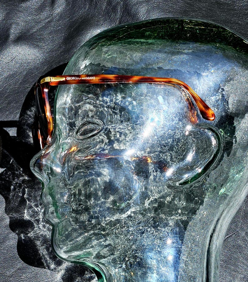May include: Tortoiseshell-patterned eyeglasses with a brown and gold frame. The temple arm features the text GIORGIO ARMANI in gold lettering. The glasses are positioned on a clear glass mannequin head, showcasing the curved temple design.