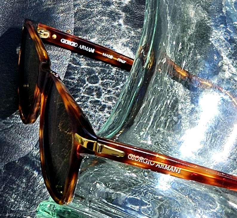 May include: Tortoiseshell-patterned sunglasses with dark lenses and gold-tone metal accents. The inner temple arm features the text Giorgio Armani and Made in Italy, while the outer temple arm displays the Giorgio Armani brand name in gold lettering.