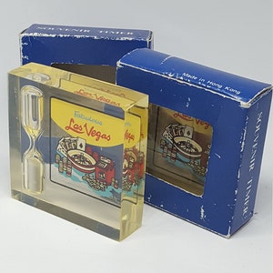 May include: A clear acrylic hourglass with a yellow base and a colorful image of a Las Vegas casino scene. The hourglass is in a blue box with the text "Fabulous Las Vegas" and "Souvenir Timer" printed on it.
