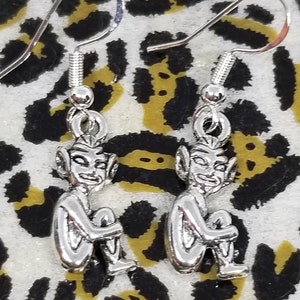 May include: A pair of silver-colored earrings featuring small, seated goblin figures. The goblins have pointed ears and are holding their knees. The earrings have a hook closure.
