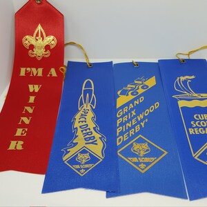 May include: Four blue and one red satin ribbons with gold lettering and images. The red ribbon says "I'M A WINNER". The blue ribbons say "SPACE DERBY", "GRAND PRIX PINEWOOD DERBY", and "CUB SCOUT REGATTA".