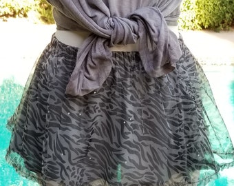May include: A black and gray zebra print tulle skirt with a ruffled hem. The skirt is worn over a gray t-shirt tied at the waist.