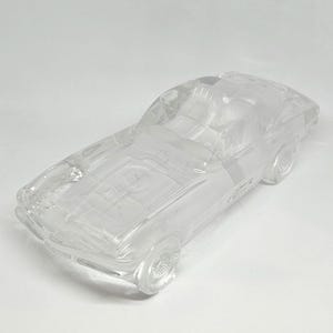 May include: A clear glass car figurine with detailed etching. The car has visible wheels, a windshield, and interior detailing. The transparent material allows light to pass through, highlighting the car's design. The figurine is a decorative item.
