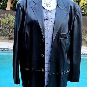 Pu&ograve; includere: Un blazer in pelle nera con revers dentellato, tre bottoni frontali e una tasca sul petto. Sotto, un top grigio con scollo a V, un sottile motivo a ragnatela e una collana color argento con un ciondolo a forma di teschio.