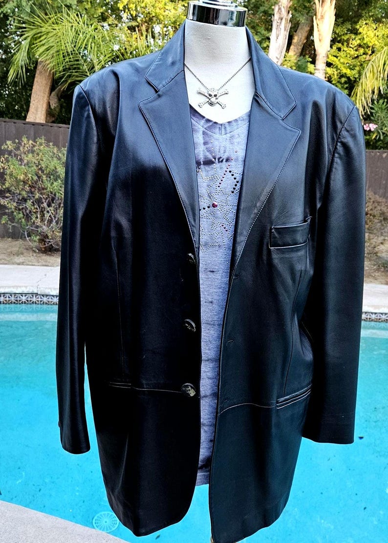 Pu&ograve; includere: Un blazer in pelle nera con revers dentellato, tre bottoni frontali e una tasca sul petto. Sotto, un top grigio con scollo a V, un sottile motivo a ragnatela e una collana color argento con un ciondolo a forma di teschio.