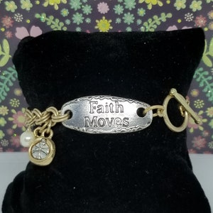 May include: A gold and silver bracelet with a charm that says "Faith Moves". The charm is oval shaped and has a textured surface. The bracelet has a toggle clasp.