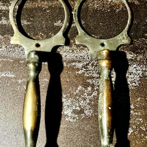 May include: Two antique brass door handles. Each handle features a decorative top with a circular opening and a long, cylindrical grip. The handles have a weathered, aged appearance, suggesting they are vintage or reclaimed.