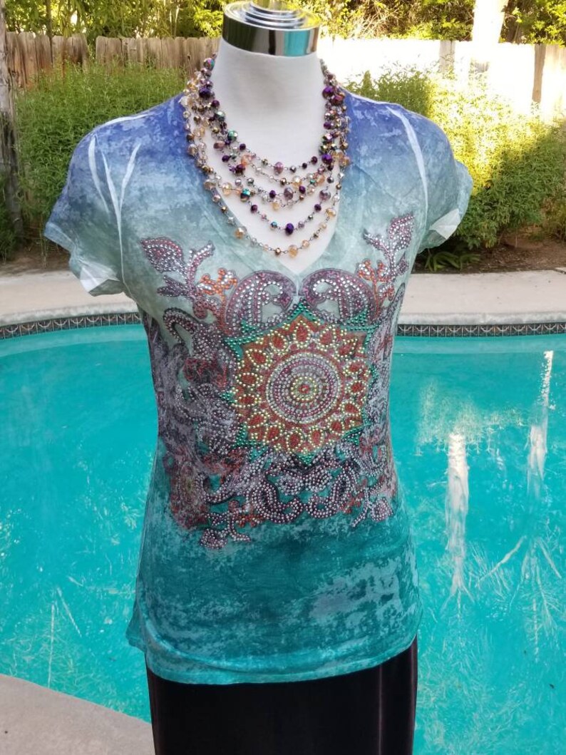May include: A teal blue, green, and purple t-shirt with a beaded design and a multi-strand beaded necklace.